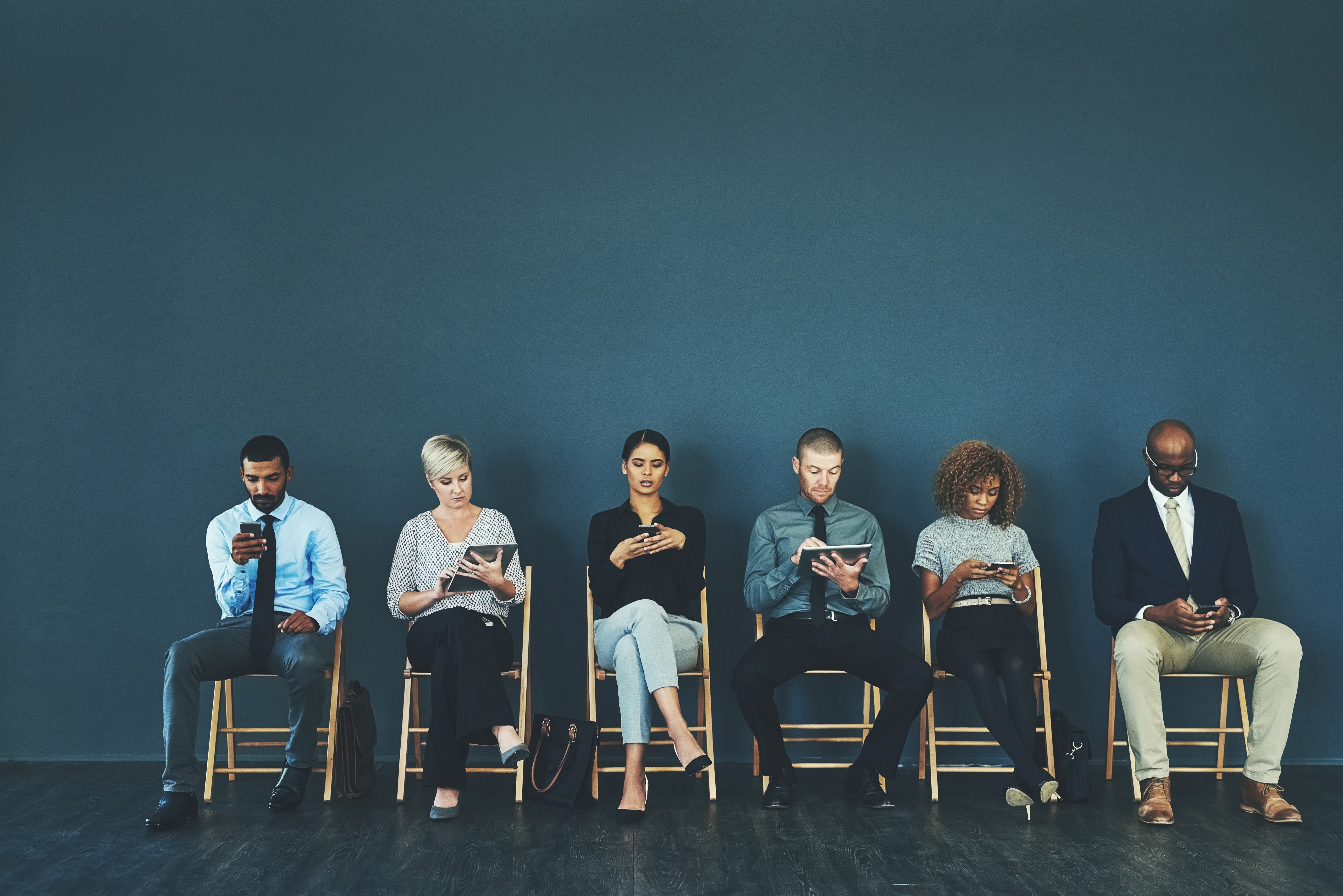 Various women and men sitting down on chairs lined up against a wall, each on their phone